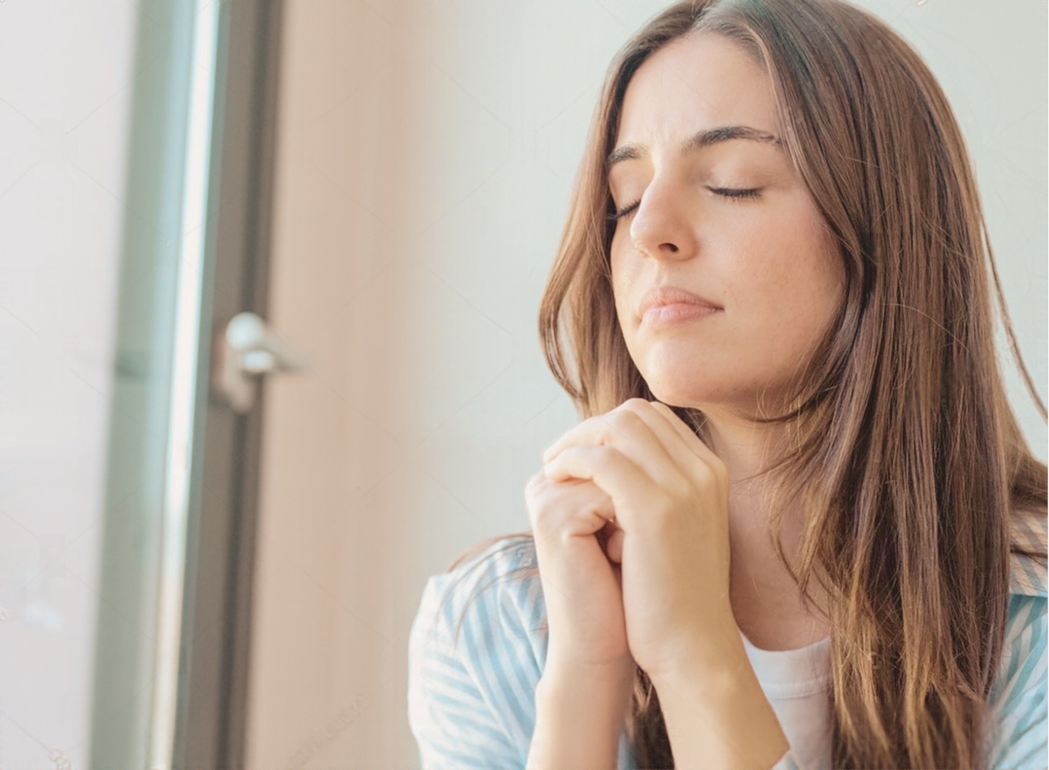 A woman with her eyes closed and hands clasped in prayer beside a bright window